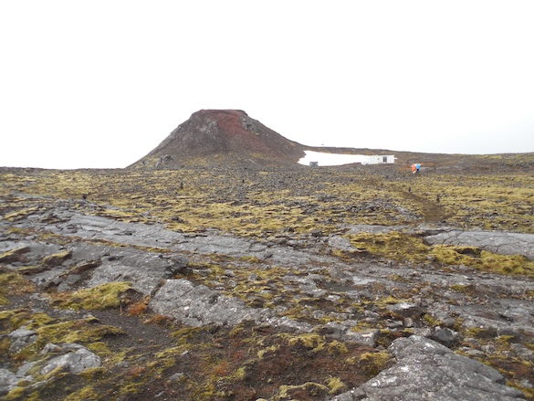 View of Thrihnukagigur Cone and the Inside the Volcano hut (at the base to the right) from the surrounding lava fields. Photo Credit: Ellie Was